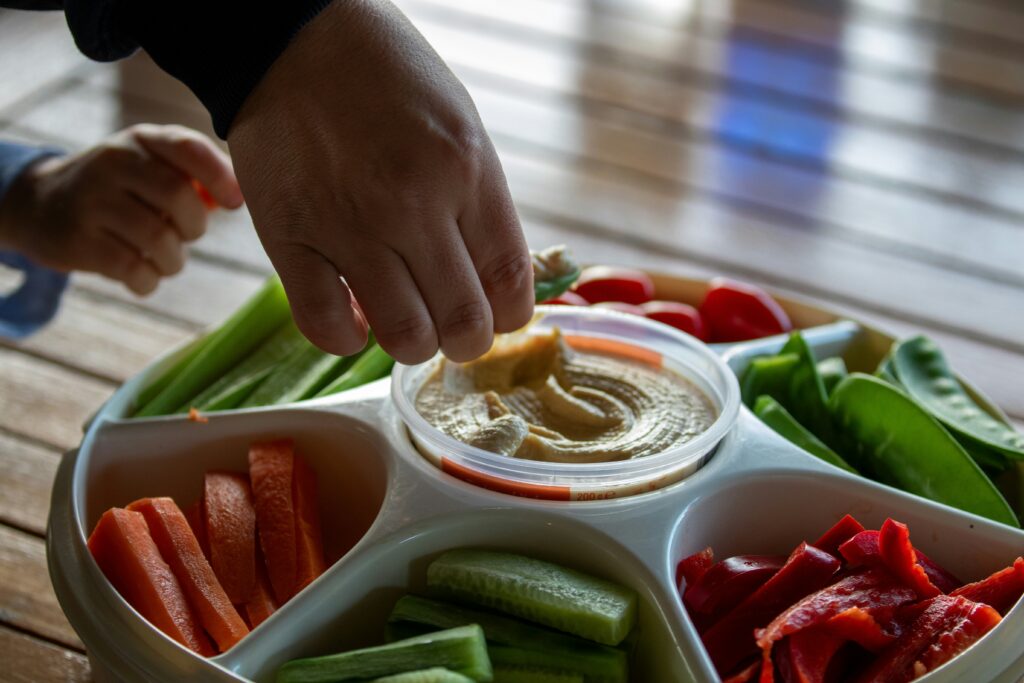 A wooden board with a bowl of creamy hummus surrounded by colorful vegetable